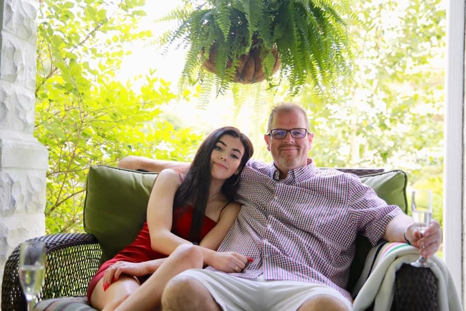 Emily sits (left) with her dad (right) on a couch, appearing to be outside with lots of greenery in the background.